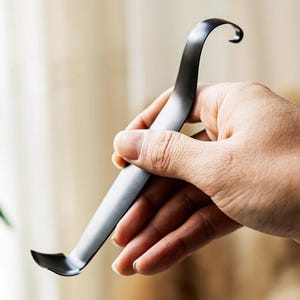 May include: A dark gray metal tool with a curved handle and a hooked end. The tool has a flat, angled blade at the opposite end. The tool is held in a hand against a blurred background.