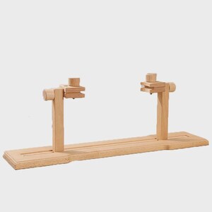 May include: A wooden embroidery hoop stand. The stand is made of light-colored wood and features a rectangular base with two vertical supports. Each support has an adjustable clamp for holding an embroidery hoop. The stand is designed to hold the hoop in place.