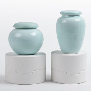 May include: Two light blue ceramic urns, one round and one tall, each with a matching lid. The urns are displayed on white, cylindrical pedestals. The background is plain white.