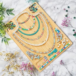 May include: A wooden jewelry design board with various beaded necklaces in different colors and sizes. The necklaces are arranged in a semi-circle, with measurements from 14 inches to 34 inches. Scattered beads and floral accents complete the display.
