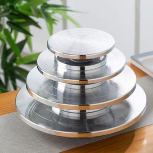 May include: A stack of four silver metal rotating cake stands of varying sizes. Each stand has a circular design with concentric rings. The stands are on a wooden table with a gray runner, with green foliage in the background.