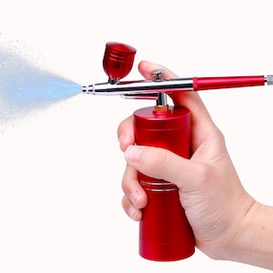 May include: A red airbrush tool with a silver nozzle and accents, spraying a fine mist of blue liquid. The airbrush is held in a hand against a white background. The tool has a cylindrical base and a trigger.