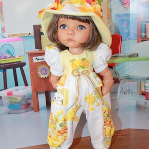 May include: A doll wearing a yellow and white floral outfit, including a hat, top, and pants. The doll has blue eyes and brown hair. A small white teddy bear is in the doll's pocket. The doll is wearing yellow shoes.