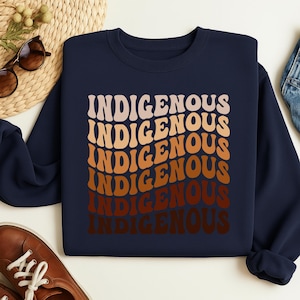 Könnte beinhalten: Marineblaues Sweatshirt mit dem Wort "INDIGENOUS" in einem Retro-Schriftzug, wiederholt in einem Farbverlauf aus Beige, Orange und Braun. Das Sweatshirt wird auf einer weißen Oberfläche präsentiert, mit Sonnenbrille und einer gewebten Matte im Hintergrund.