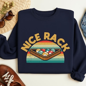 Puede incluir: Sudadera azul marino con un diseño retro que presenta un taco de billar y las palabras "NICE RACK" en amarillo. El diseño incluye un triángulo de bolas de billar en un taco de madera, sobre un fondo a rayas.