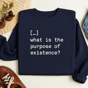 May include: A navy blue sweatshirt featuring the white text "[-] what is the purpose of existence?". The sweatshirt has a crew neck and is a solid colour. The image also shows sunglasses, a woven mat, and brown shoes.