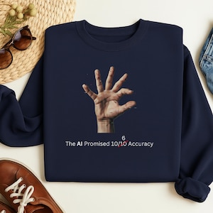 Op de afbeelding: Marineblauwe sweatshirt met een hand en de tekst "The AI Promised 10/10 Accuracy" met het cijfer 6 doorgestreept. De hand staat in het midden van de sweatshirt.