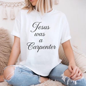 May include: White t-shirt with the text "Jesus was a Carpenter" in a black script font. The shirt has short sleeves and a relaxed fit. The model is wearing light blue jeans.
