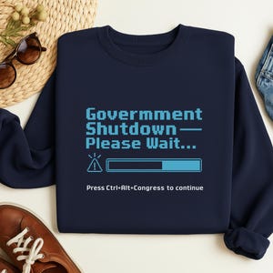 Puede incluir: Sudadera azul marino con el texto "Government Shutdown - Please Wait..." en fuente pixelada. Debajo, una barra de carga y la frase "Press Ctrl+Alt+Congress to continue."
