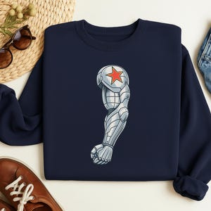 May include: Navy blue sweatshirt featuring a detailed illustration of a silver and gray robotic arm with a red star on the shoulder. The design is centered on the front of the sweatshirt.