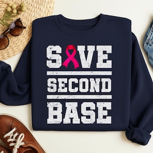 Peut inclure: Sweat-shirt bleu marine avec l'inscription "SAVE SECOND BASE" en lettres blanches usées. Un ruban rose est intégré au mot "SAVE". Ce vêtement décontracté est adapté au quotidien.