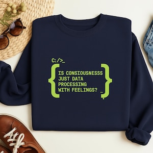 May include: A navy blue sweatshirt featuring the text "IS CONSIOUSNESSS JUST DATA PROCESSING WITH FEELINGS?" in green. The design includes code-like brackets and the prompt "C:/_>" in green.