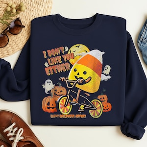 Op de afbeelding: Marineblauwe sweatshirt met een Halloween-thema. Het ontwerp toont een snoepmaïskarakter op een fiets, omringd door pompoenen, geesten en de tekst "I DON'T LIKE YOU EITHER" en "HAPPY HALLOWEEN ANYWAY".