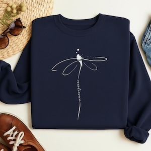 Può includere: Felpa blu navy con un disegno di libellula bianca. La parola "resilience" è scritta verticalmente sotto. La felpa è di colore uniforme e sembra essere realizzata in un materiale morbido. Un top casual.