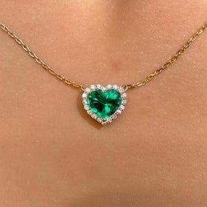 Può includere: Una collana a catena dorata con un ciondolo a forma di cuore caratterizzato da una pietra centrale verde brillante. Il cuore è circondato da un delicato alone di piccole pietre chiare e scintillanti, conferendo un aspetto raffinato al gioiello.