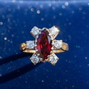 Puede incluir: Un anillo de oro con una gran gema roja oscura de forma ovalada en el centro, rodeada de gemas claras más pequeñas. El anillo se presenta sobre un fondo azul oscuro, creando un contraste llamativo. La banda del anillo tiene un diseño detallado.