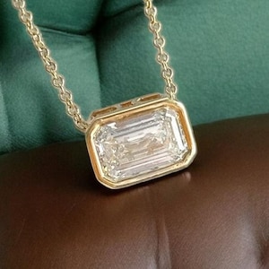 Puede incluir: Un collar de oro con un gran colgante rectangular de piedra preciosa transparente. El colgante está engastado en un bisel de oro y cuelga de una delicada cadena de oro. El fondo es de tela verde oscuro.