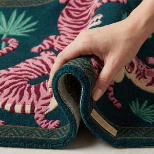 May include: A rolled-up teal rug with a pink tiger design. The rug features a detailed pattern of tigers, palm leaves, and a geometric border. The rug is made of a soft, textured material.