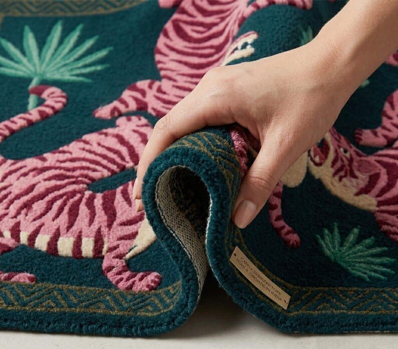 May include: A rolled-up teal rug with a pink tiger design. The rug features a detailed pattern of tigers, palm leaves, and a geometric border. The rug is made of a soft, textured material.