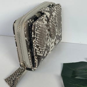 May include: A bi-fold wallet with a snakeskin pattern in shades of grey, white, and black. The wallet has a silver zip closure and a matching pull tab. The interior is not visible.