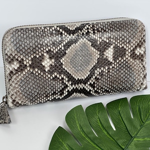 Pode incluir: Uma carteira retangular com um padrão de pele de cobra em tons de cinza, preto e branco. A carteira tem fecho de correr e um pequeno borla. O fundo é branco com uma folha verde.
