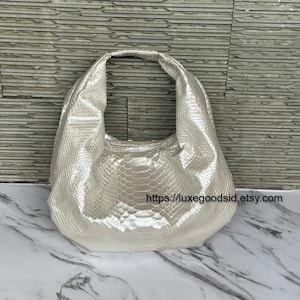 Peut inclure: Un sac à main argenté texturé, de forme incurvée, avec une seule bandoulière. Le sac présente un motif peau de serpent et est présenté sur une surface en marbre. L'URL de la boutique Etsy est visible.