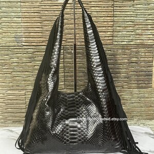 May include: A black leather shoulder bag with a snakeskin pattern and fringe detailing. The bag has long handles and a large, open design. The bag is displayed on a metal stand against a textured background. The website address is visible on the bag.