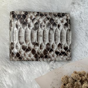 Può includere: Un portafoglio a due ante con un motivo a pelle di serpente nei toni del grigio e del marrone. Il portafoglio è rettangolare e sembra essere in pelle. La trama della pelle di serpente è chiaramente visibile.