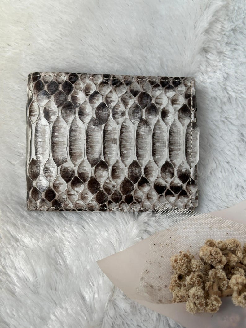 Puede incluir: Una cartera plegable con un patr&oacute;n de piel de serpiente en tonos grises y marrones. La cartera es rectangular y parece estar hecha de cuero. La textura de la piel de serpiente es claramente visible.