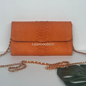 May include: An orange clutch with a chain strap. The clutch is rectangular with a flap closure and a textured, snakeskin-like pattern. The gold chain strap is draped in front of the bag. The text "LuxeGoodsID" is printed on the front.