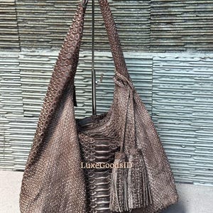 Könnte beinhalten: Braune Schultertasche aus Leder mit strukturierter Reptilienmusterung. Die Tasche hat ein lässiges Design, einen langen Riemen und eine Quaste. Sie wird auf einem Metallständer präsentiert.