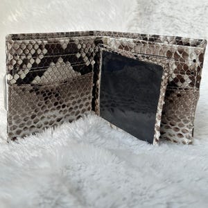Puede incluir: Cartera bi-fold abierta con un estampado de piel de serpiente en tonos marr&oacute;n, gris y blanco. La cartera tiene m&uacute;ltiples ranuras para tarjetas y una ventana de identificaci&oacute;n transparente. El interior es de color negro s&oacute;lido.