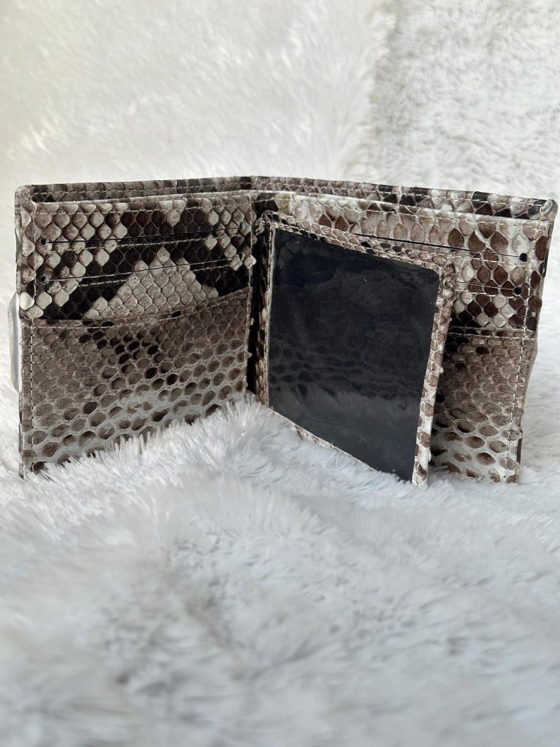 Puede incluir: Cartera bi-fold abierta con un estampado de piel de serpiente en tonos marr&oacute;n, gris y blanco. La cartera tiene m&uacute;ltiples ranuras para tarjetas y una ventana de identificaci&oacute;n transparente. El interior es de color negro s&oacute;lido.