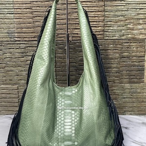 May include: A large, sage green leather hobo bag with a snakeskin pattern. The bag features long black fringe on the sides and a wide shoulder strap. The bag is displayed on a metal stand against a textured background.
