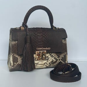 May include: A brown and cream snakeskin handbag with a top handle and a detachable shoulder strap. The bag features a gold-colored clasp and a decorative tassel. The text "LuxeGoodsID" is visible on the bag.