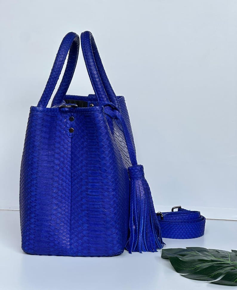 May include: A vibrant blue tote bag with matching handles and a detachable shoulder strap. The bag features a textured, snakeskin-like pattern and a decorative tassel. The bag is a stylish accessory.