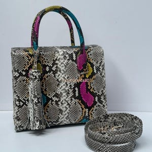 Op de afbeelding: Een vierkante handtas met een slangenprint in grijs, zwart en wit. De handvatten zijn veelkleurig, met turquoise, roze en gele accenten. Een bijpassende schouderriem ligt opgerold naast de tas. Een kwast hangt aan de handvat.