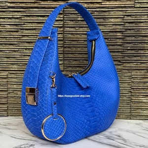 May include: A bright blue hobo bag with a textured snakeskin pattern, featuring a single top handle, a gold-toned metal clasp, and a circular gold charm hanging from a clip. The bag has a zipper closure and displays the text https://luxegoodsid.etsy.com.