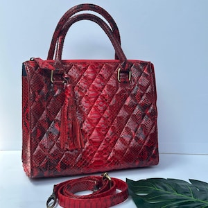 Op de afbeelding: Een rood-zwarte gewatteerde handtas met een bijpassende schouderriem. De tas heeft een handvat aan de bovenkant en een decoratieve kwast. De afmetingen van de tas zijn ongeveer 25 cm breed, 20 cm hoog en 10 cm diep.