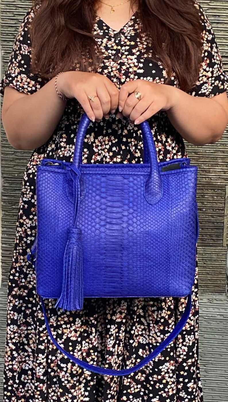 May include: A vibrant blue handbag with a textured snakeskin pattern. The bag features a top handle, a detachable shoulder strap, and a decorative tassel. The bag's design is rectangular, with a structured silhouette.
