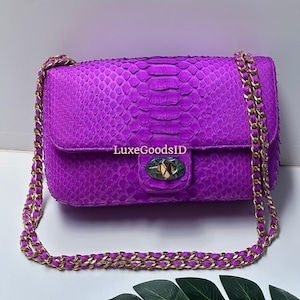 Può includere: Una vivace borsa viola con una tracolla a catena. La borsa presenta un motivo strutturato e squamoso e una chiusura dorata. La tracolla è color oro. Il testo "LuxeGoodsID" è visibile sulla borsa.