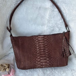 以下が含まれることがあります： 茶色のヘビ革模様のハンドバッグ。バッグにはショルダーストラップとタッセルが付いています。バッグは長方形で、ファスナー開閉式です。質感のある素材で作られています。