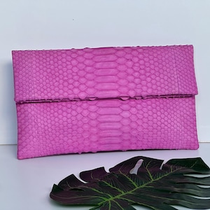 Può includere: Una pochette rosa acceso con un motivo a pelle di serpente strutturato. La borsa rettangolare ha una chiusura con patta. La borsa è realizzata in materiale di pelle di serpente. Una foglia verde scuro è in primo piano.