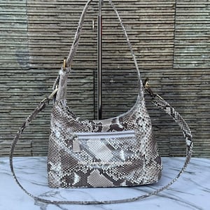 Puede incluir: Un bolso de hombro estilo hobo con un patrón de piel de serpiente en tonos gris, marrón y blanco. El bolso incluye un asa superior, una correa de hombro desmontable con herrajes en tono latón y un bolsillo horizontal con cremallera al frente.