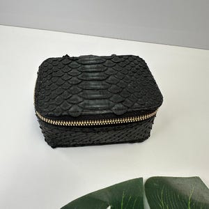 Könnte beinhalten: Kleine, schwarze, rechteckige Box mit einem strukturierten, schuppenartigen Muster. Die Box hat einen goldfarbenen Reißverschluss. Der Artikel ist wahrscheinlich eine Schmuckschatulle oder ein kleiner Aufbewahrungsbehälter.