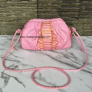 May include: Pink clutch bag with a snakeskin pattern and a gold-coloured centre panel. The bag has a braided pink strap and a gold-coloured clasp. The bag is on a white surface.