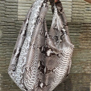 May include: A large, soft handbag with a snakeskin pattern in shades of gray, brown, and white. The bag is held up by a hand, showcasing its shape and the texture of the material. The bag's design is casual and stylish.