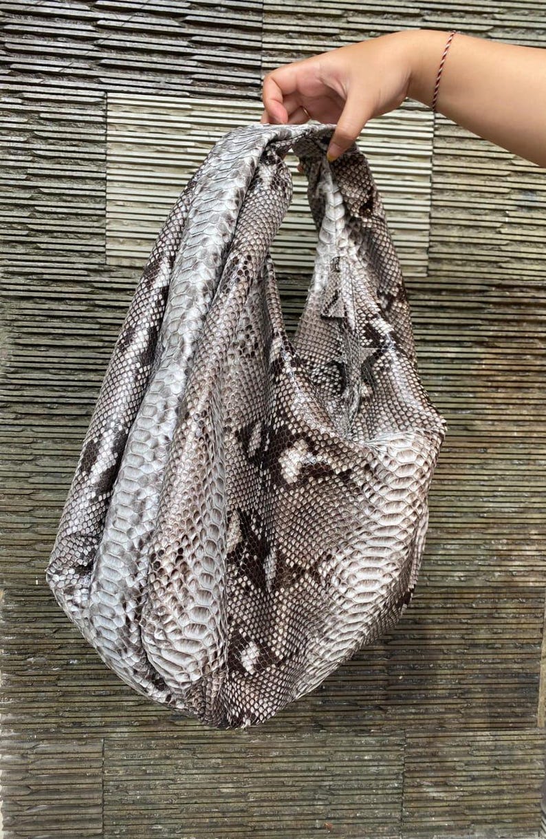 May include: A large, soft handbag with a snakeskin pattern in shades of gray, brown, and white. The bag is held up by a hand, showcasing its shape and the texture of the material. The bag's design is casual and stylish.