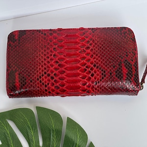 Könnte beinhalten: Eine rote Python-Haut-Geldbörse mit Reißverschluss. Die Geldbörse hat eine rechteckige Form und eine strukturierte Oberfläche. Die Farbe ist ein leuchtendes Rot mit dunkleren roten und schwarzen Akzenten.