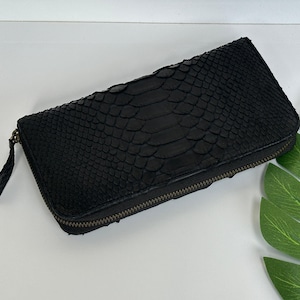 Könnte beinhalten: Eine schwarze, rechteckige Geldbörse mit einem strukturierten, schlangenlederartigen Muster. Die Geldbörse hat einen Reißverschluss und eine kleine Lasche. Ein grünes Blatt ist im Hintergrund.